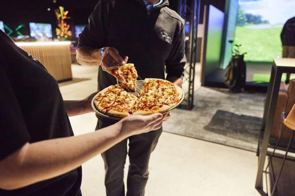 People sharing pizza at X-Golf venue with simulator bays in background