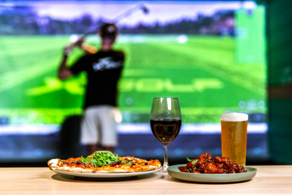 Food and drinks on table with X-Golf simulator gameplay in background