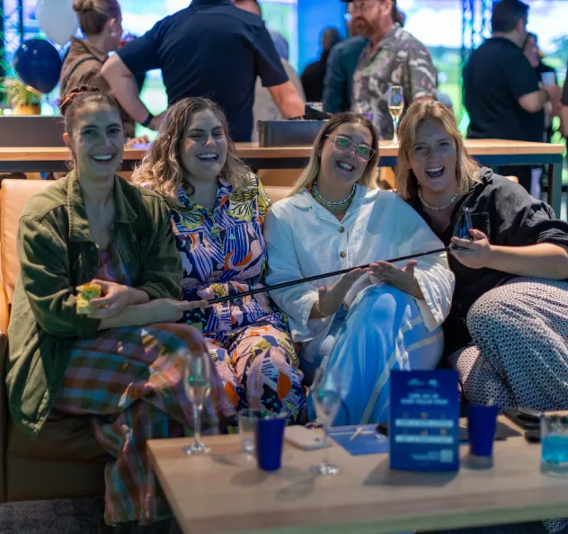 Four women sitting together on a couch, smiling and enjoying drinks at an indoor sports venue lounge.